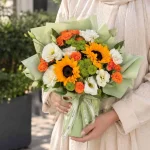 دسته گل آفتابگردان و رز نارنجی آویدا با گل‌های سفید و سبز و کاغذپیچ سبز روشن
